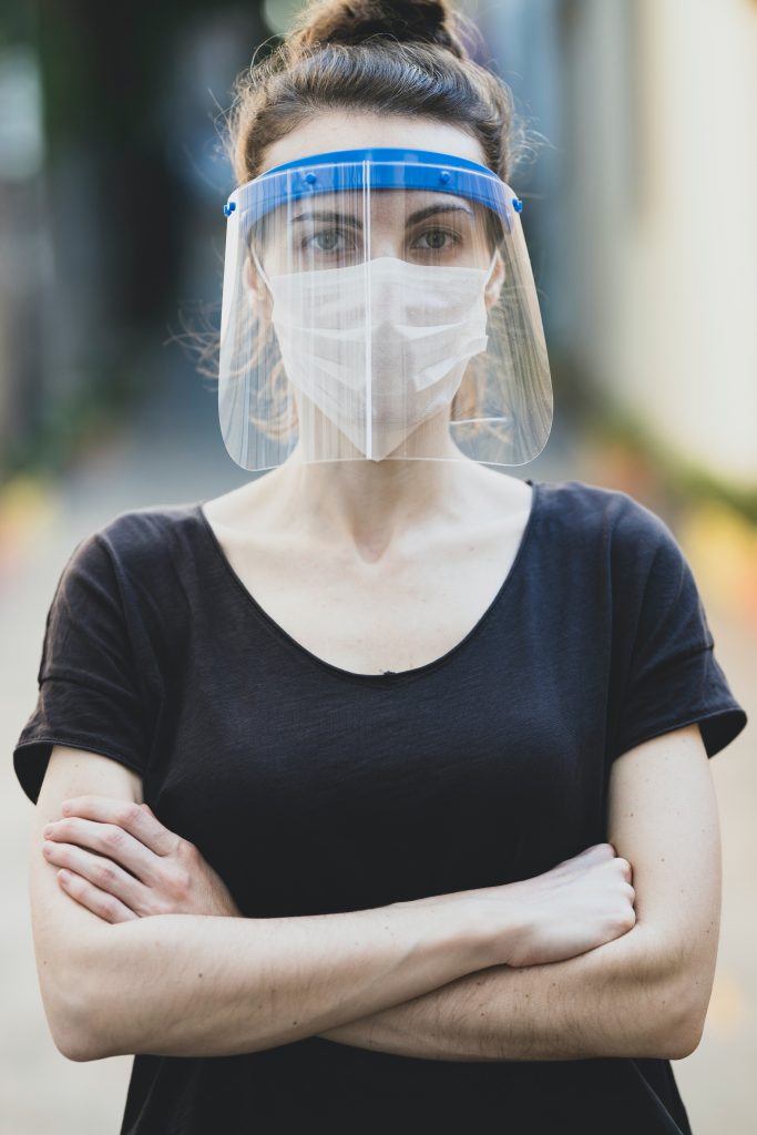 Faire tomber les masques, vraiment ? Une jeune femme portant masque et visière ne plexiglas. elle nous regarde droit dans les yeux et croise les bras