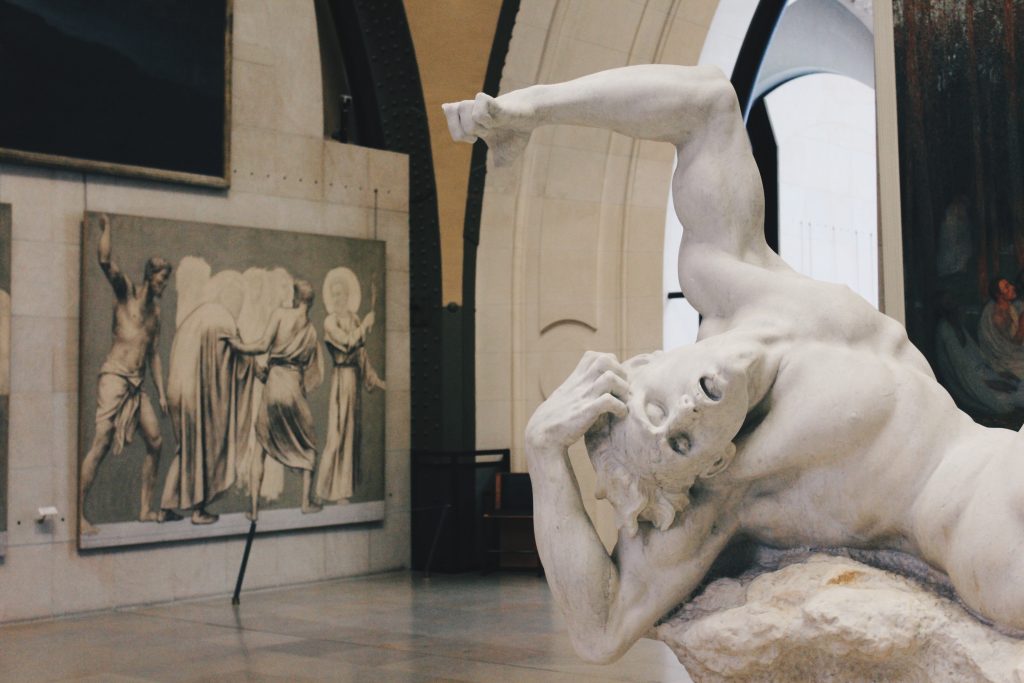 dans un musée, une sculpture d'un homme couché qui se tord de douleur et crie la bouche ouverte. au fond, un grand tableau avec une scène évoquant la souffrance